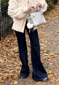 Krémově texturovaný svetr, béžový kardigan, tmavé rozšířené džíny, šedá kabelka s přívěskem a dekorativním bambulkou, stojící na cestě pokryté listím.