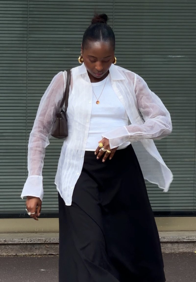 Frau trägt eine durchsichtige weiße Bluse über einem weißen Top, schwarzen Rock, goldene Creolen, goldene Ringe und eine braune Schultertasche, steht im Freien.