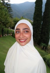 Hiyab de color crema claro, hecho de una tela suave, con un brillo sutil. El fondo presenta follaje verde y montañas distantes. Ojos azules, brillantes y claros.