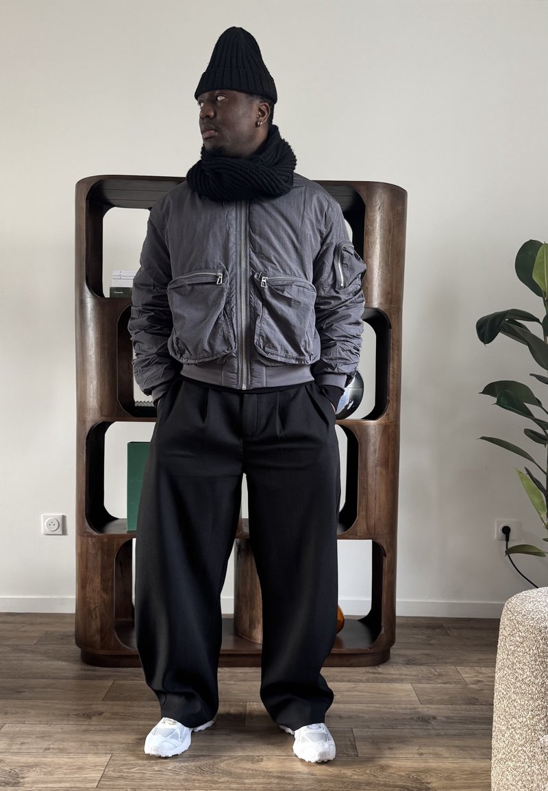 Homme portant un bonnet noir, une écharpe noire, une veste grise zippée avec de grandes poches, un pantalon large noir et des baskets blanches, debout à l'intérieur.