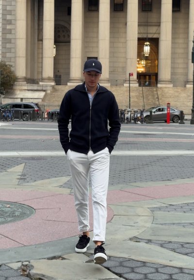 Chaqueta de cremallera azul marino sobre una camisa azul clara, pantalones blancos y zapatillas negras. De pie sobre un pavimento texturizado con arquitectura clásica de fondo.