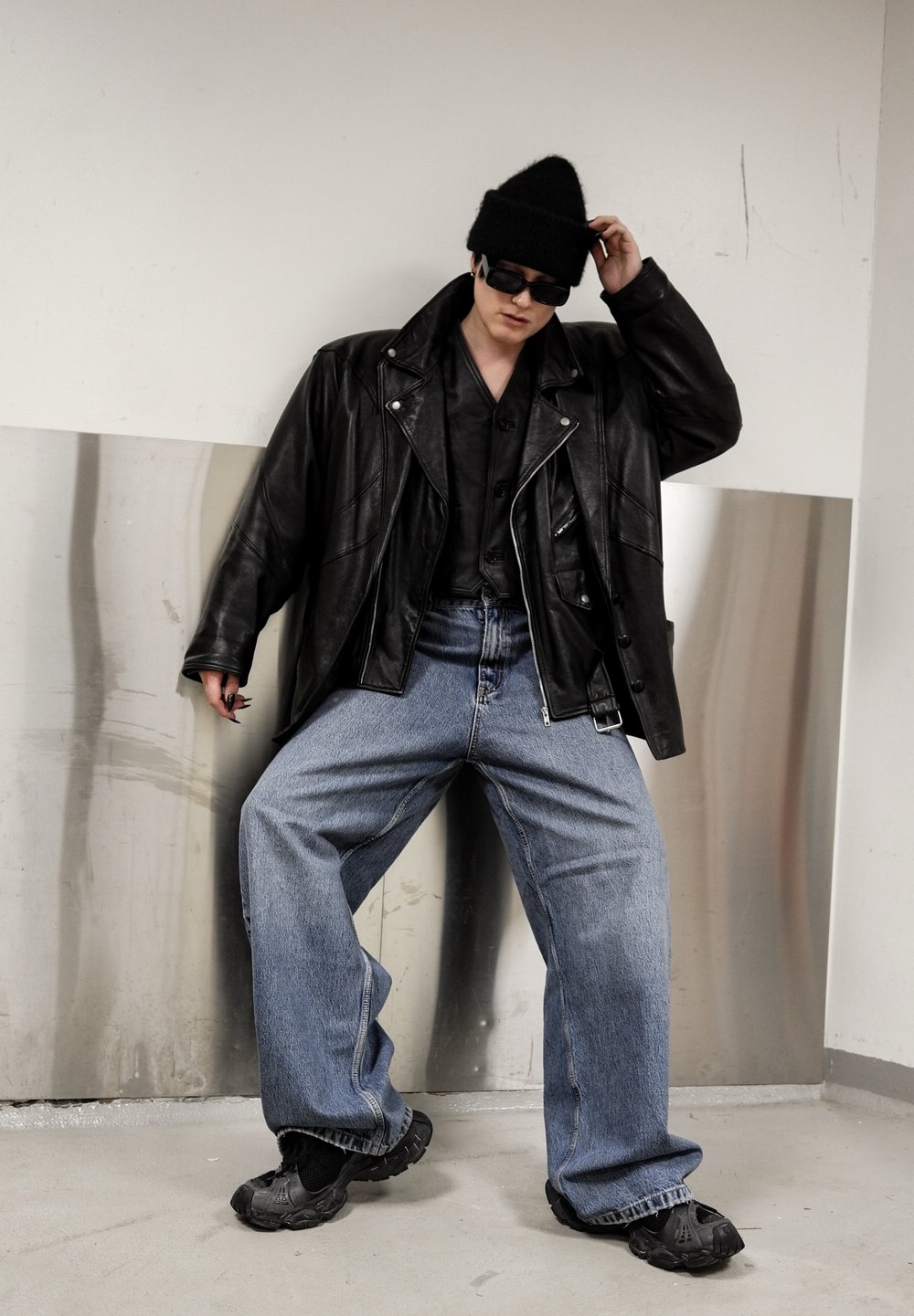 Veste surdimensionnée en cuir noir, chemise noire à boutons, jean ample en denim bleu, bonnet noir, lunettes de soleil et chaussures noires au design unique.