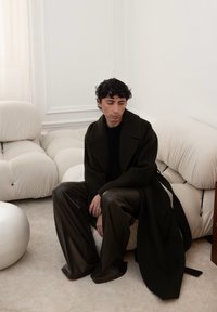 Dunkelgrüner langer Mantel über einem schwarzen Oberteil, kombiniert mit weiten braunen Lederhosen. Lockige Haare, sitzend gegen ein hellfarbiges, plüschiges Sofa.