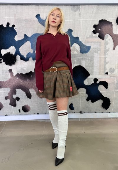 Camisa de manga larga burdeos combinada con una falda plisada a cuadros, cinturón marrón, calcetas blancas hasta la rodilla con rayas rojas y tacones negros de punta.