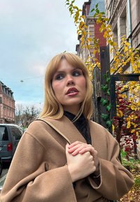 Manteau enveloppant en laine marron avec des manches larges, chemise noire en dessous, mains croisées sur la poitrine. Texture douce, lumière naturelle, feuilles d'automne en arrière-plan.