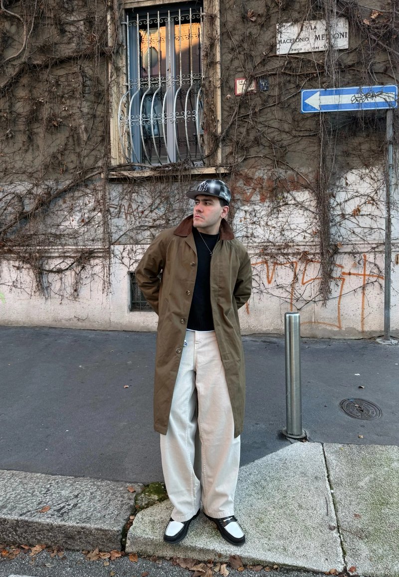 Homme portant un long manteau brun, un pantalon large blanc, une chemise noire et une casquette noire, debout dans la rue près d'un mur couvert de lierre avec une fenêtre grillagée et des graffitis.