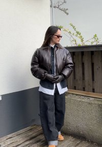 Rjav usnjen pufferjakna z elastičnimi manšetami, prekrita nad belo srajco in črnimi denim hlačami. Oblečeni črni rokavici in veliki sončna očala.