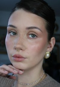 Jeune personne à la peau claire et tachetée de rousseur, portant un anneau de septum en or, de grandes boucles d'oreilles en perles, deux colliers et un pull en tricot, reposant le menton sur la main.