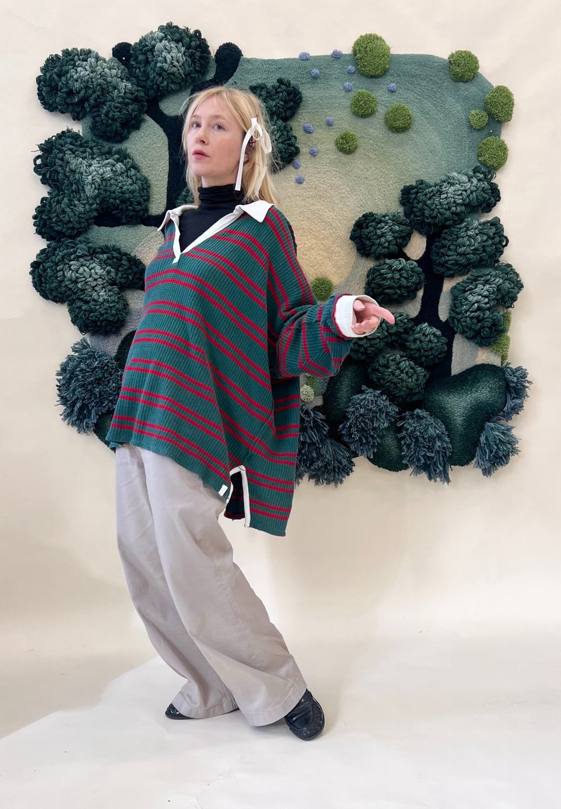 Jeune femme dans un pull surdimensionné rayé vert et rouge et un pantalon beige pose devant une œuvre d'art murale texturée verte et bleue avec des formes d'arbres.