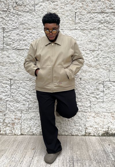 Chaqueta beige con cremallera y cuello, dos bolsillos y un pequeño logo. Pantalones negros y zapatos grises completan el look, frente a una pared texturizada.