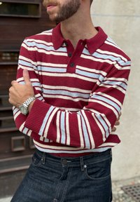 Punasest ja kreemikas triibuline pullover kraega, millel on sinised detailid ja kolm musta nööpi, kombineeritud tumedate teksapükstega. Tekstuuriga kootud kangas.