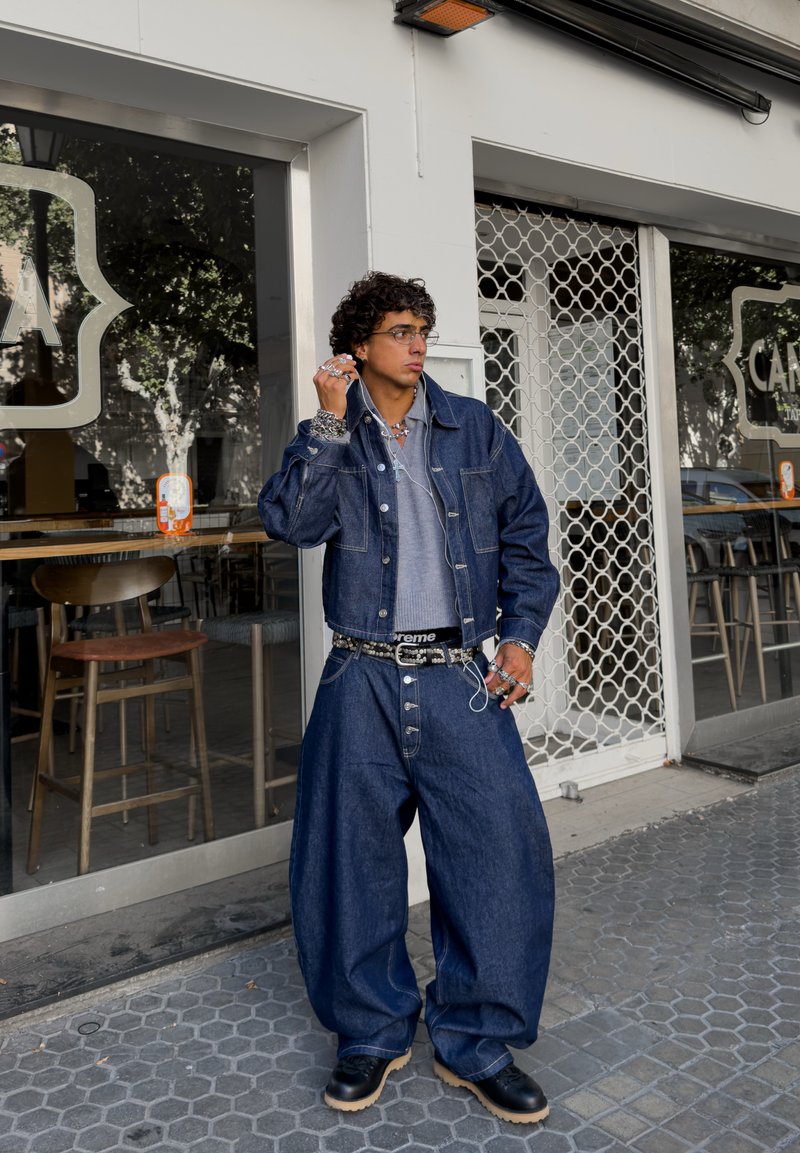 Joven con cabello rizado que lleva una chaqueta de mezclilla oversized y pantalones, de pie frente a un café con unos auriculares en la mano.