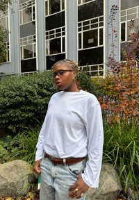 Fehér hosszú ujjú ing, világoskék farmer, barna öv. Kiegészítők: arany fülbevalók és gyűrűk. Természetes háttér zöld növényzettel.