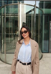 Camisa de colarinho azul claro, casaco oversized bege e calças bege claras com um cinto preto. Óculos de sol redondos e escuros e cabelo longo.