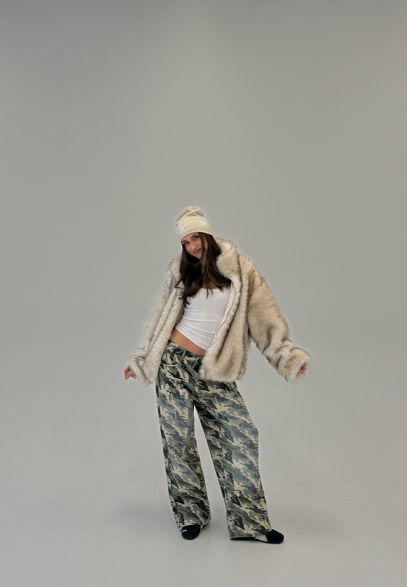 Nuori nainen, jolla on valkoinen pipo, beigen värinen tekoturkistakki, valkoinen toppi ja maastohousut, seisoo yksivärisen harmaan taustan edessä.