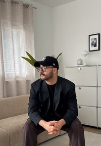 Crna dvoredna blazer jakna preko crne košulje, sivo hlače i crna kapa. Nosi se prozirne naočale. Unutarnji ambijent s dekorativnom biljkom i namještajem.