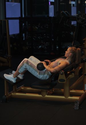 Mujer en ropa de entrenamiento acostada en un banco inclinado, sosteniendo una mancuerna sobre el estómago en un gimnasio con poca luz, rodeada de máquinas de peso.