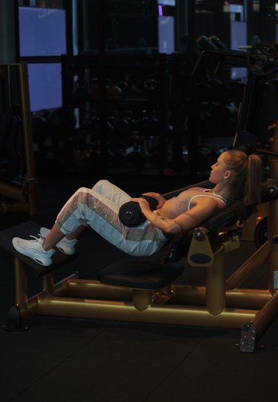 Mujer en ropa de entrenamiento acostada en un banco inclinado, sosteniendo una mancuerna sobre el estómago en un gimnasio con poca luz, rodeada de máquinas de peso.