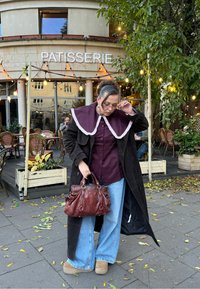 Barna kordbársony kabát túlméretezett szilvaszín gallérral, bordó ing, bő szárú kék farmer és barna kézitáska. Patiszéria háttér lombos növényzettel.