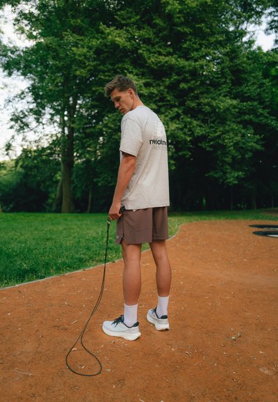 Camiseta gris con logotipo negro, pantalones cortos marrones, zapatillas deportivas blancas, sosteniendo una correa negra de perro, de pie en un camino de tierra en un área con hierba.