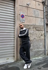 Άνδρας με μαύρη φόρμα Adidas και λευκά αθλητικά παπούτσια ακουμπά σε τοίχο κοντά σε μεταλλικό ρολό και πινακίδα απαγόρευσης στάθμευσης σε αστικό περιβάλλον.