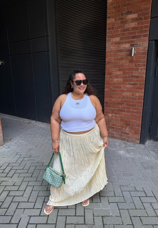 Femme portant un débardeur blanc, jupe plissée crème, lunettes de soleil, tenant un sac à main vert, debout sur un trottoir pavé près de murs en briques noires et rouges.