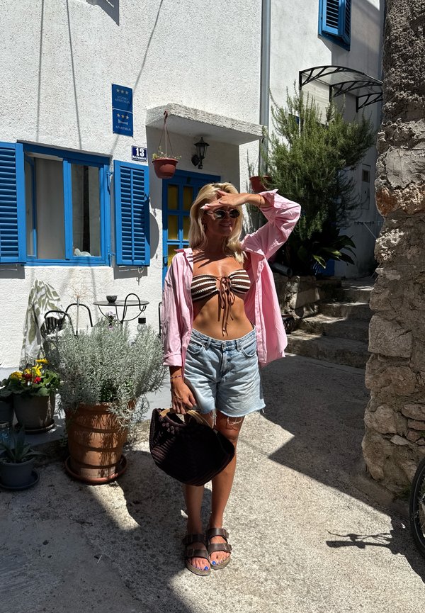 Femme en lunettes de soleil et vêtements d'été décontractés, se protégeant les yeux du soleil, debout dans une rue ensoleillée près d'un bâtiment blanc avec des volets bleus et des plantes en pot.