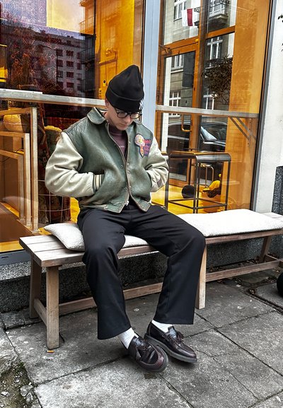 Chaqueta de varsity de cuero verde y tela crema con parches, pantalones oscuros y zapatos negros. Sentado en un banco de madera fuera de un café.