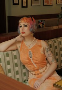 Oranje jurk met plooidetail en een ovale uitsnijding. Beaded kettingriem, parelketting en kleurrijk kort haar met highlights.