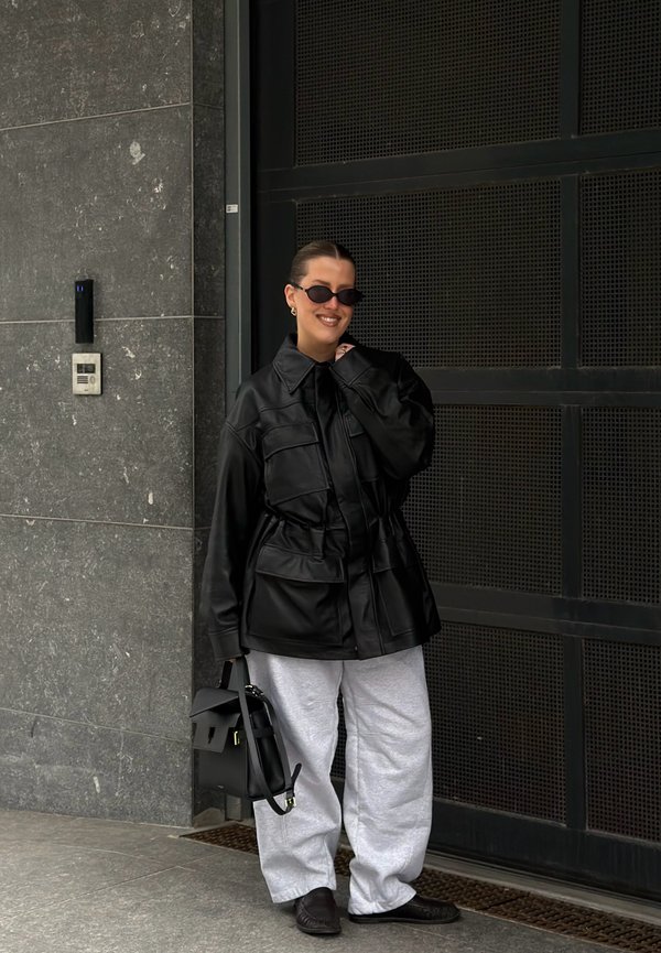 Žena nosi preveliku crnu kožnu jaknu, svijetlo sive trenirke, crne cipele, sunčane naočale te drži crnu ručnu torbu, stoji pored tamnih metalnih vrata.