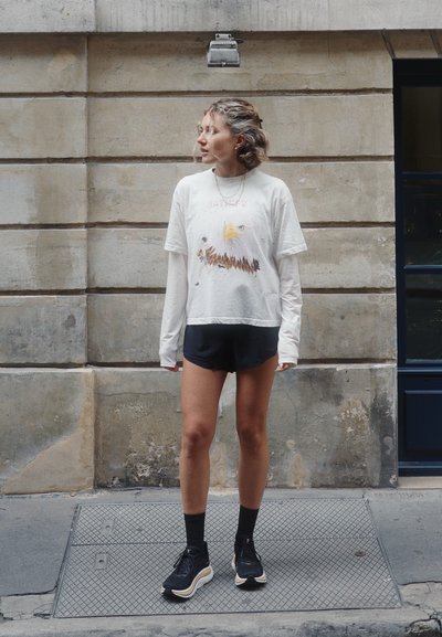 Mujer con cabello rubio, camiseta blanca de manga larga de capas con diseño de águila, pantalones cortos negros, calcetas negras y zapatillas, de pie en una calle de la ciudad.