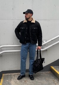 Veste à col en fausse peau de mouton noire portée sur un pull noir, jean bleu clair, chaussures noires et sac noir. En arrière-plan, un mur gris et des escaliers.