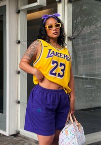 Yellow basketball jersey featuring "Lakers" and the number 23, teamed with purple shorts. Accessories consist of a white bag with a vibrant pattern and sunglasses.