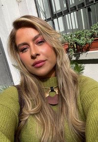 Maglione verde a maglia con un motivo testurizzato, collana in oro e marrone a strati, capelli biondi lunghi e sfondo illuminato delicatamente con piante.