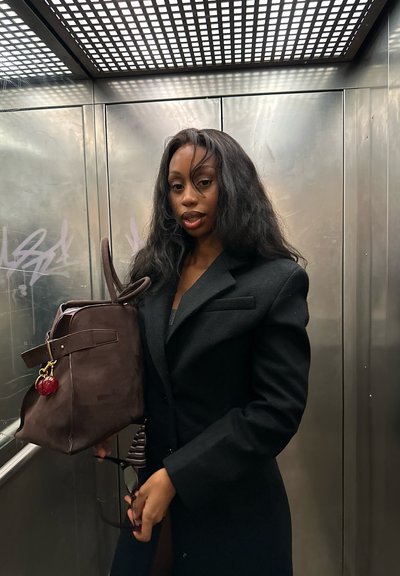 Mujer con cabello largo y oscuro, vestida con un abrigo negro, que sostiene un bolso marrón y gafas de sol dentro de un ascensor metálico con grafitis en las paredes.