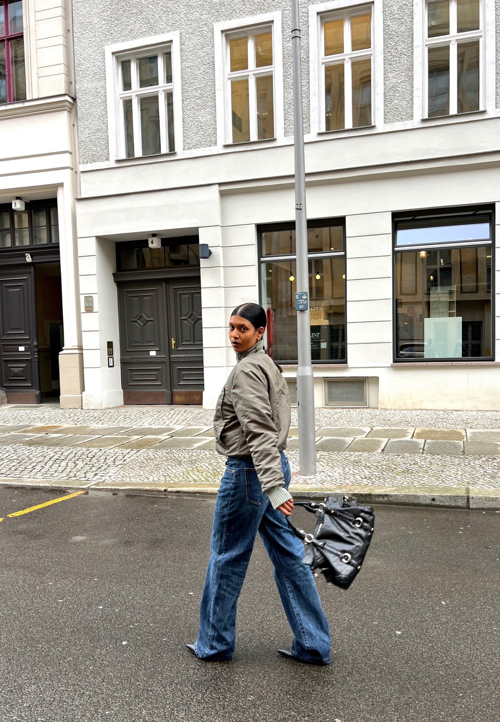 Graue Bomberjacke, lockere blaue Jeans, schwarze Handtasche mit silbernem Zubehör, läuft auf einer Kopfsteinpflasterstraße in der Nähe eines Stadtgebäudes.