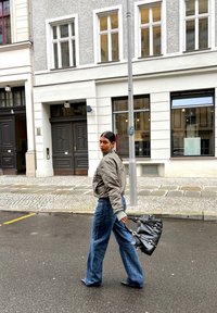 Graue Bomberjacke, lockere blaue Jeans, schwarze Handtasche mit silbernem Zubehör, läuft auf einer Kopfsteinpflasterstraße in der Nähe eines Stadtgebäudes.