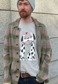Chemise à carreaux verte et rose portée sur un t-shirt gris orné de deux Dalmatiens de dessin animé avec un cœur, sur fond d'un mur texturé.
