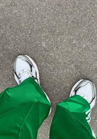 Valged spordijalatsid võrguga ja hallide detailidega, roheliste läikivate võistlusdressipükstega, asetatud tekstureeritud hallile pinnale.