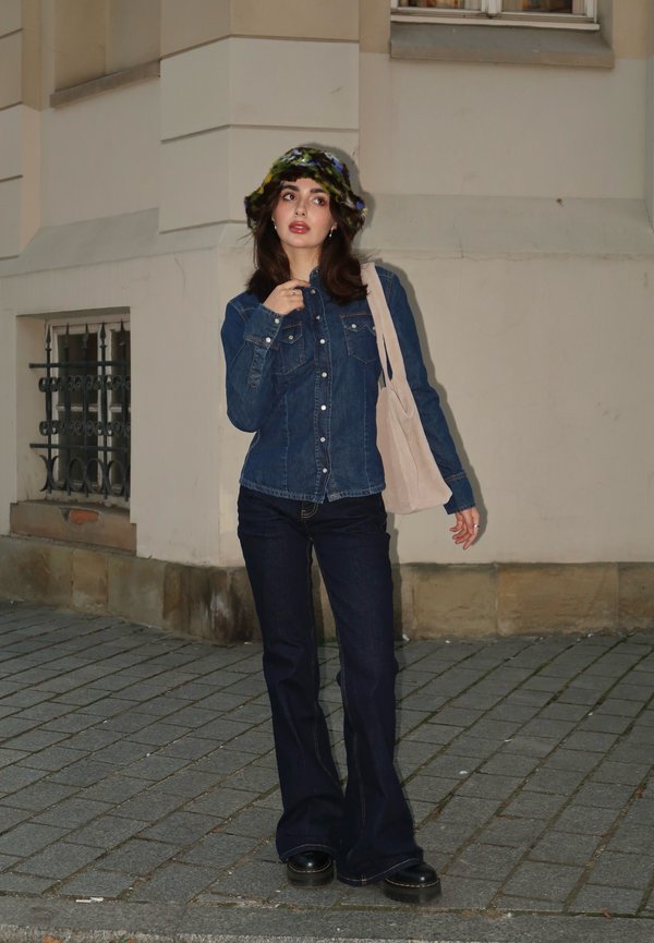 Giacca di denim con bottoni a pressione, abbinata a jeans scuri a zampa d'elefante e un cappello a secchiello lavorato a maglia multicolore. Borsa in tela a tracolla in mano.