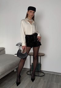 Bijela satenska košulja s gumbima, crne šorts s sekvencama, prozirne tajice, crne stiletto cipele i crna torba s okruglom ručkom.