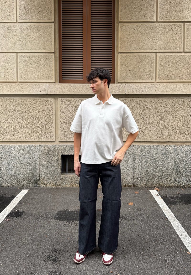 Jeune homme debout sur l'asphalte devant un mur beige texturé avec une fenêtre à volets marron, portant un polo blanc, un jean large foncé et des baskets.