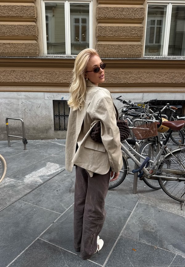 Une femme blonde portant des lunettes de soleil et un blazer beige regarde derrière elle sur un trottoir en ville, à côté de vélos garés et d’un bâtiment en pierre.
