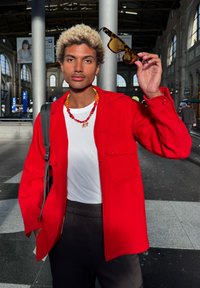 Personne aux cheveux blonds bouclés, vêtue d'une veste rouge et d'un collier de perles, tenant des lunettes de soleil en écaille de tortue à l'intérieur d'une grande gare.