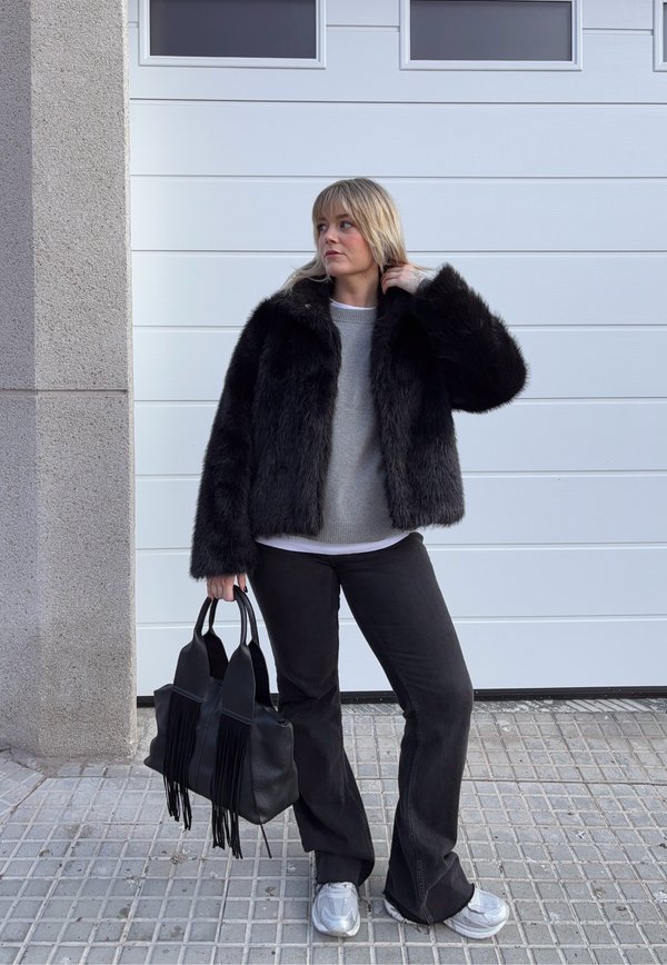 Femeie purtând o geacă neagră de blană artificială, pulover gri, pantaloni negri și adidași argintii, ținând o geantă neagră cu franjuri, stând pe o pavimentare cu plăci.