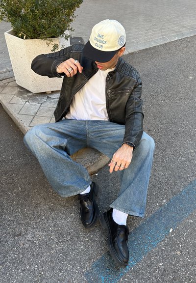 Chaqueta de cuero negra, camiseta blanca, jeans de pierna ancha de color azul claro, zapatos negros y una gorra crema con letras azules, sentado contra un jardinera.