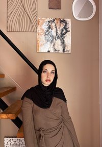 Καφέ φαρδύ φόρεμα με υφή, μακρυά μανίκια και δεμένη μέση, συνοδευόμενο από μαύρη μαντίλα. Ξύλινες σκάλες και αφηρημένη τοιχογραφία στο παρασκήνιο.