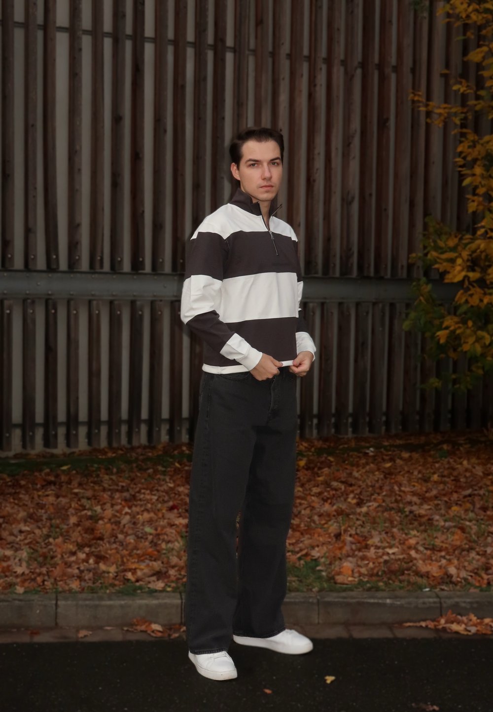 Hvit og mørkebrun stripete genser med glidelås i halsen, kombinert med svarte bukser med vide ben og hvite joggesko, foran en trebakgrunn.