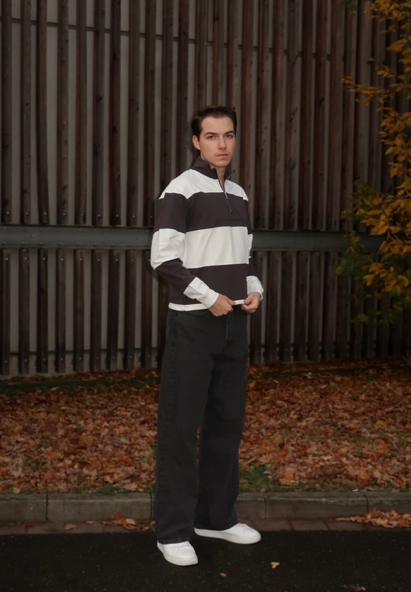 Hvit og mørkebrun stripete genser med glidelås i halsen, kombinert med svarte bukser med vide ben og hvite joggesko, foran en trebakgrunn.