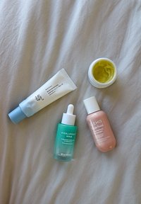 Quattro prodotti per la cura della pelle disposti su un tessuto chiaro: un tubetto bianco con tappo blu, una bottiglia con contagocce verde, un flacone per crema rosa e un barattolo per crema giallo.